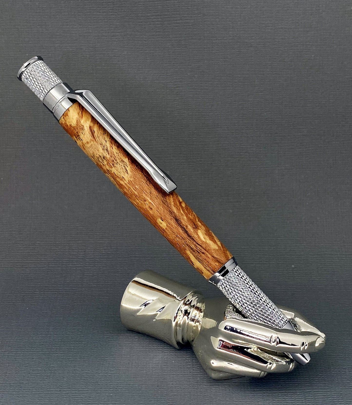 A right hand shaped metal base holding a handturned Mottled Oak wood pen as you would hold it to write with.