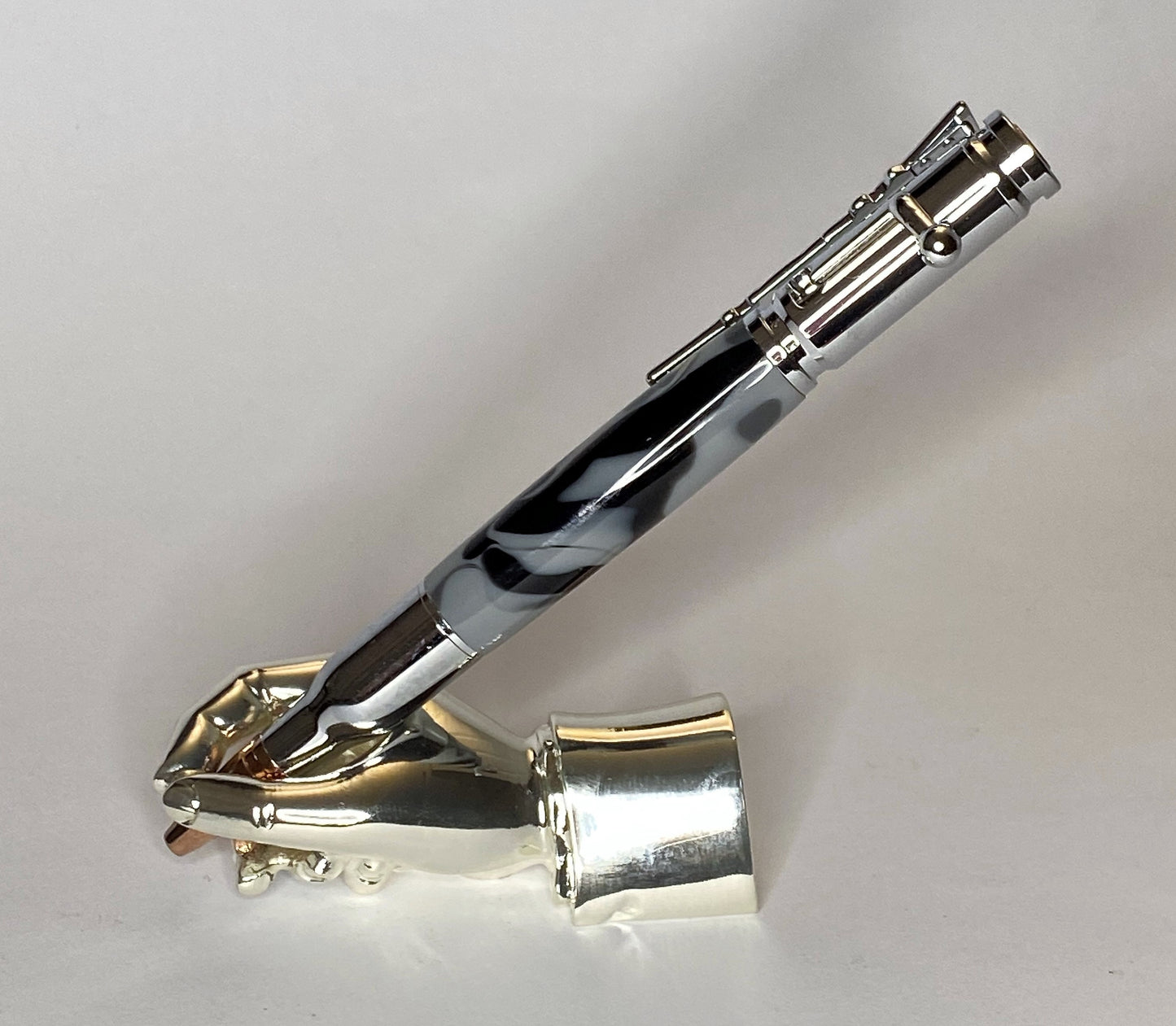 A right hand shaped metal base holding a handturned Grey cammo acrylic Bolt action pen as you would hold it to write with.