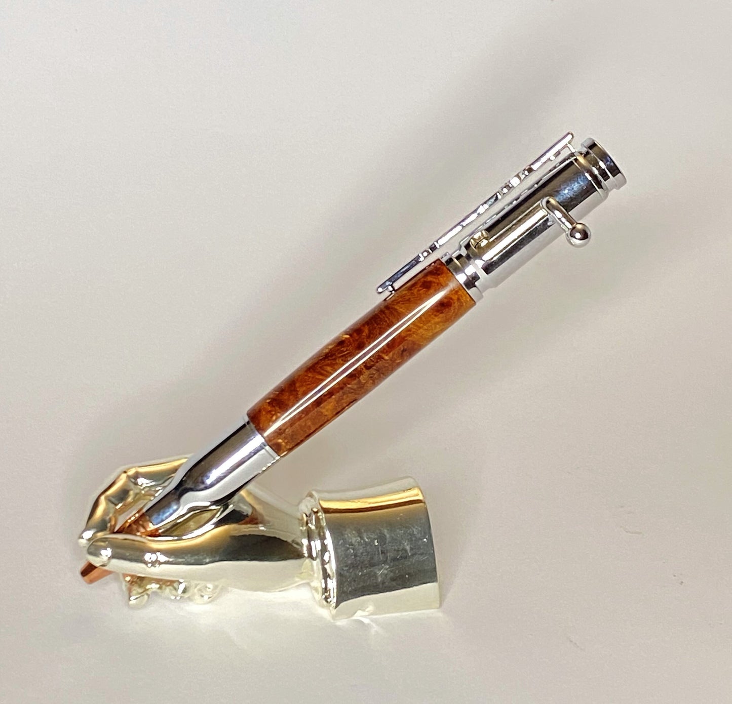 A right hand shaped metal base holding a handturned Popular Burr wood Bolt action pen as you would hold it to write with.