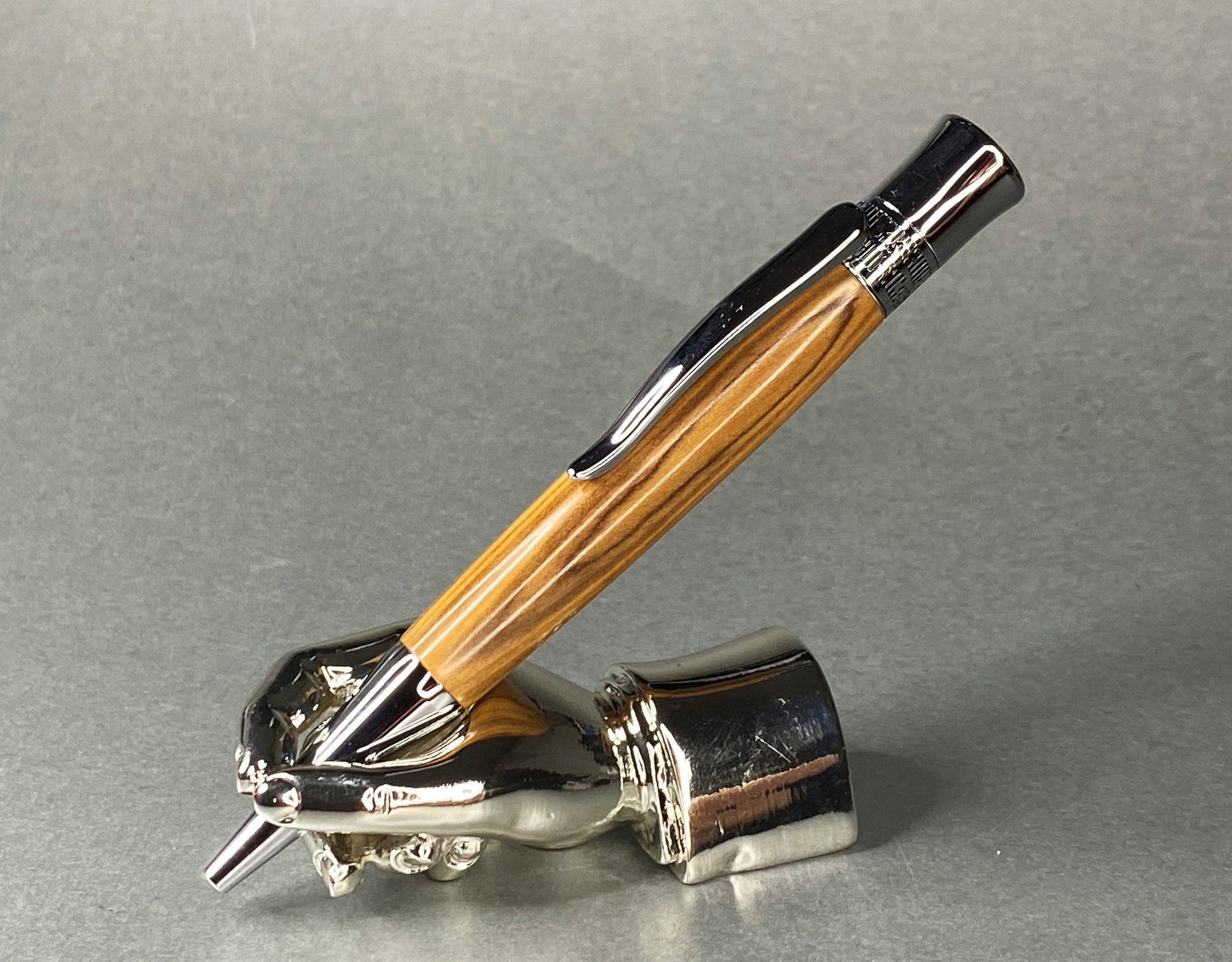 A right hand shaped metal base holding a handturned Olive Wood pen as you would hold it to write with.